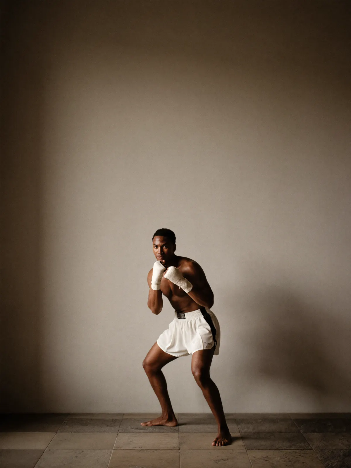 A boxer in quiet guard stance with cloth hand wraps against a plain wall
