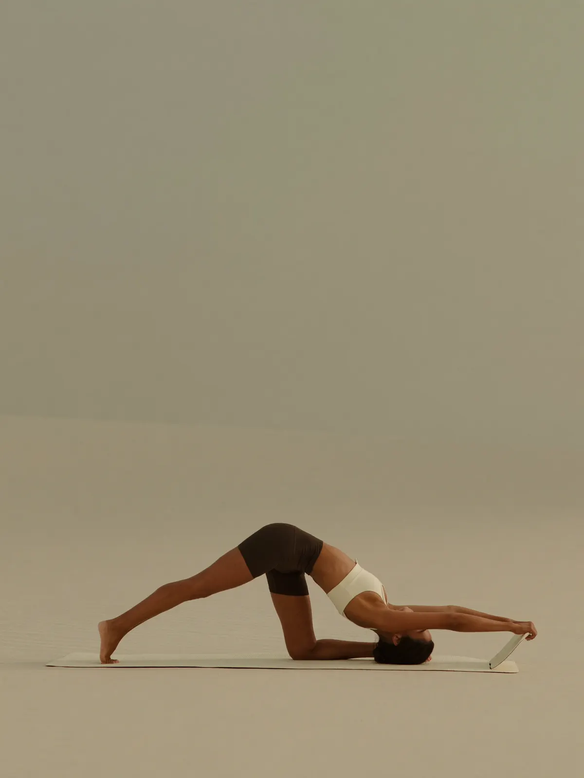 A woman holding a Pilates boat pose on a mat on pale sand under a vast pastel sky